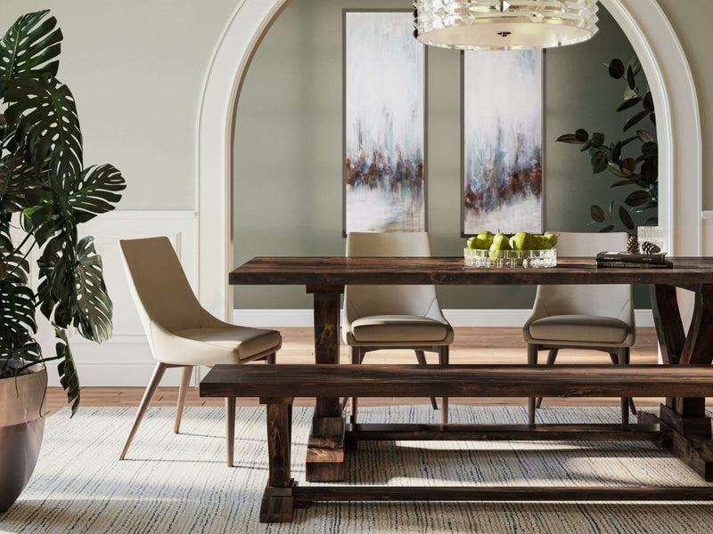 A modern dining room features a rustic wooden table with the Vera Bench in Tobacco from James+James and four beige chairs. The room is adorned with two abstract paintings on a muted green wall, a large potted plant, and a chandelier. A bowl of green apples serves as the centerpiece on the table.