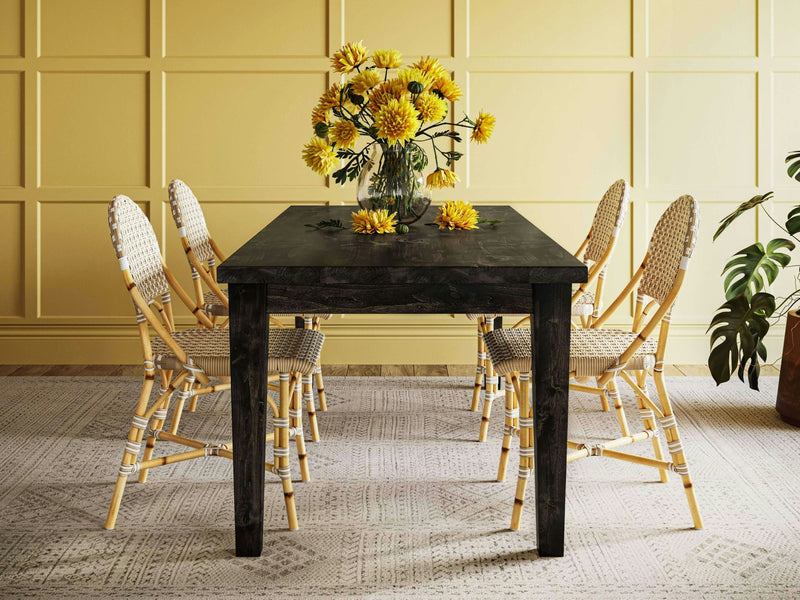 The Ava Parsons Dining Table in Charred Ember by James+James is adorned with a clear vase of yellow flowers. Surrounding the table are four wicker chairs with plaid cushions. The room features a yellow paneled wall, light-colored flooring with a patterned rug, and a potted plant.