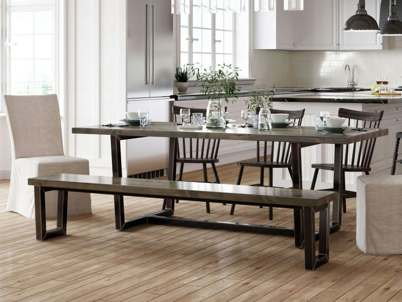 A modern, rustic dining area featuring a wooden table paired with the Trapezoid Bench in Deep Grey from James+James, along with matching chairs. The table is elegantly set with plates, glasses, and cutlery. The room showcases light-colored walls, a wooden floor, and large windows. In the background, there's a kitchen outfitted with white cabinets and pendant lights.