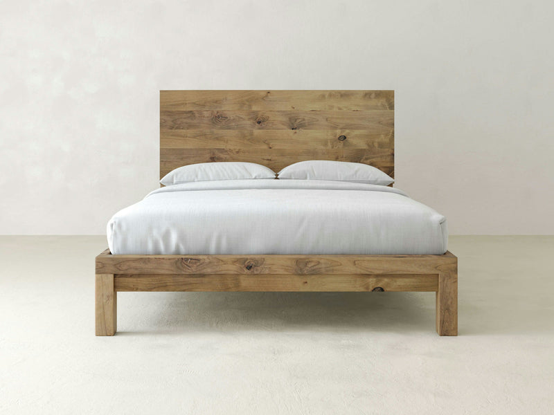 A minimalist bedroom features the Sawyer Bed in Harvest Wheat by James+James, boasting a wooden frame with a high headboard and an elegantly simple design. The bed is neatly made with white bedding and two pillows. The room follows a neutral color scheme with off-white walls, maintaining an uncluttered look without any other visible furniture or decorations.