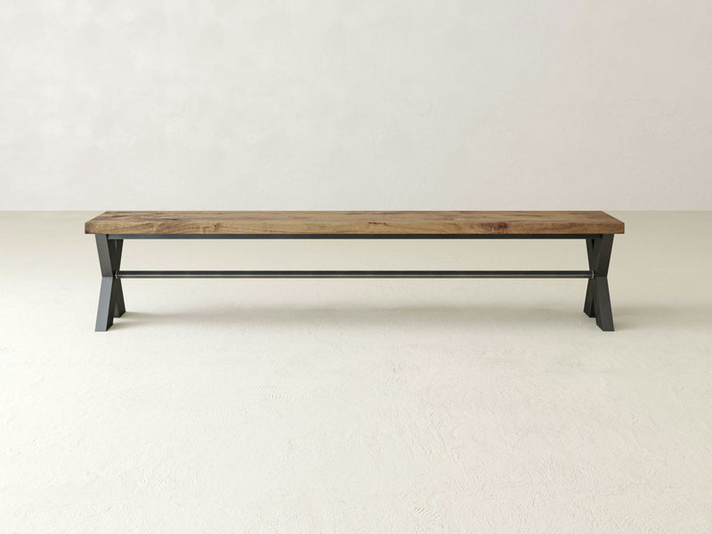 A minimalist X-Base Bench - Harvest Wheat from James+James, featuring a natural wood finish and black metal legs, is centered against a plain off-white background. The bench showcases a simple, rustic design with a flat seat and a supportive metal rod connecting the legs for added stability.