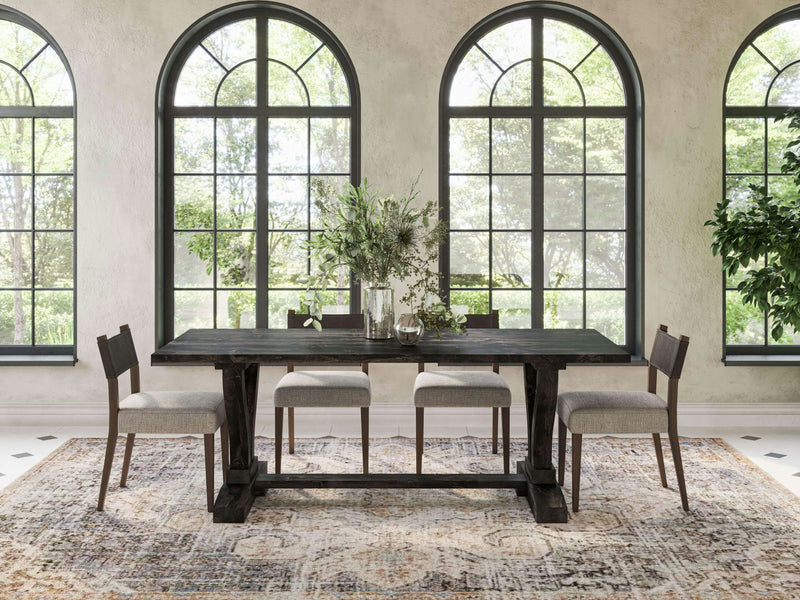 A dining area featuring the Vera Dining Table in Charred Ember by James+James, accompanied by four matching chairs, is set on a patterned rug. Behind the table are large arched windows showcasing a view of lush greenery. The table is adorned with a centerpiece of plants and decorative items. Natural bright lighting fills the room.