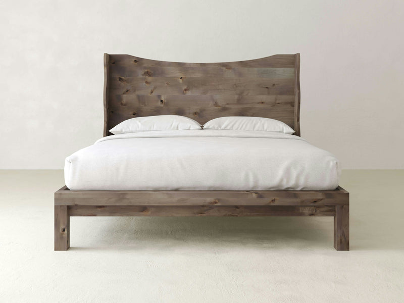 The Harlow Bed - Barn Wood by James+James is a wooden bed frame with a rustic design, featuring a tall, curved headboard and a minimalistic footboard. It is crafted from natural, light-toned wood with visible grain patterns and is dressed with white bedding, including pillows and a neatly made duvet.