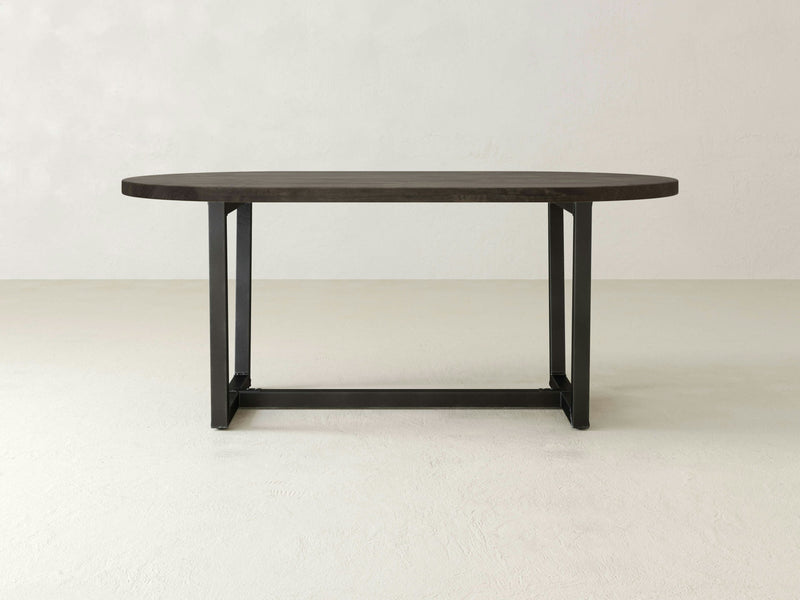 The Trapezoid Oval Dining Table - Deep Grey by James+James features a dark grey wooden oval top with a black metal base, set on a light-colored floor against a plain, light background. The minimalist design boasts clean lines and a sturdy frame, giving it a modern, sleek appearance.