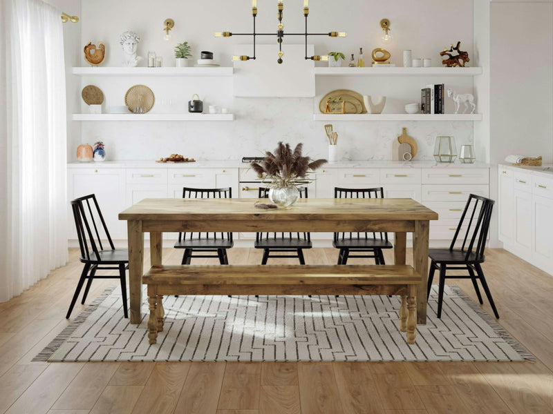 A dining room featuring a rustic Annli Bench in Harvest Wheat from James+James, paired with six black chairs around a patterned rug. The backdrop consists of white cabinets and open shelves adorned with various decor items, including plates, plants, and vases. A modern chandelier hangs above the table.