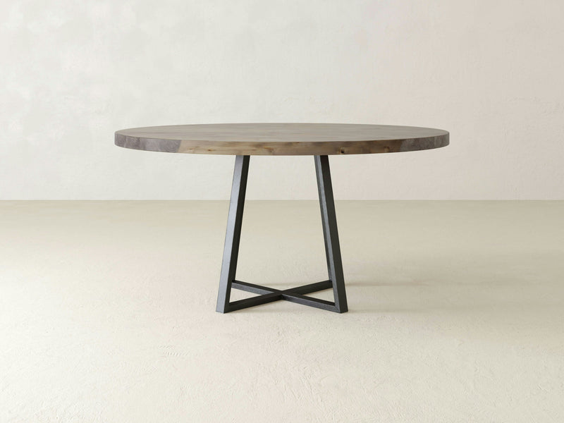 A Watson Round Dining Table - Barn Wood crafted by James+James, featuring a robust wooden tabletop paired with a contemporary metal base in a geometric design. The backdrop consists of plain, light-colored walls and flooring, enhancing the minimalist and clean aesthetic.