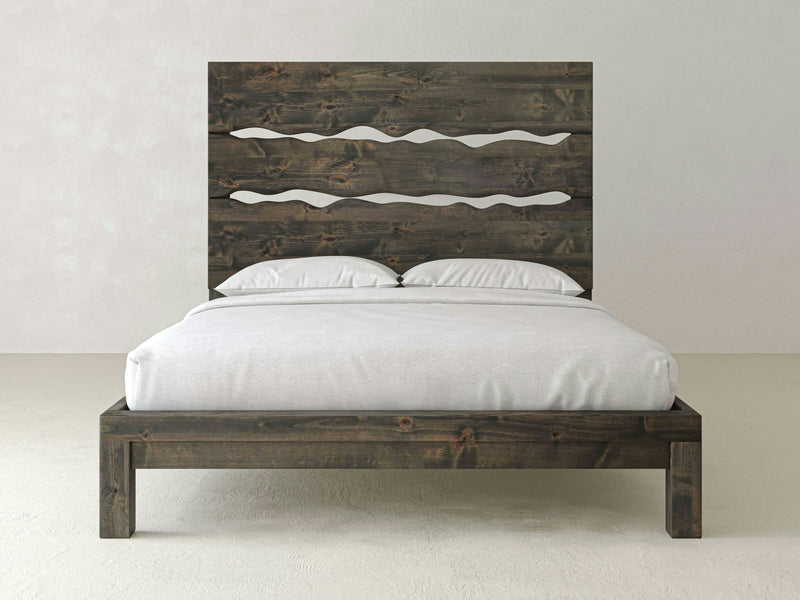 The Live Edge Bed - Deep Grey by James+James features a unique headboard design with intricate cut-out wave patterns. Crafted from dark-stained wood, this bed frame is neatly made up with crisp white bedding and two coordinating white pillows. The minimalist setting is complemented by a light, neutral-colored wall and floor.