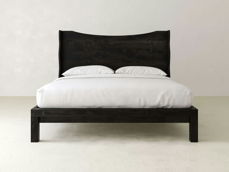 The Harlow Bed - Charred Ember by James+James features a minimalist, modern design with a dark wooden frame and headboard. This bed is neatly made with a white duvet and two white pillows, set against a plain, light-colored wall and floor. The overall look is simplistic and elegant, emphasizing clean lines and contrast.