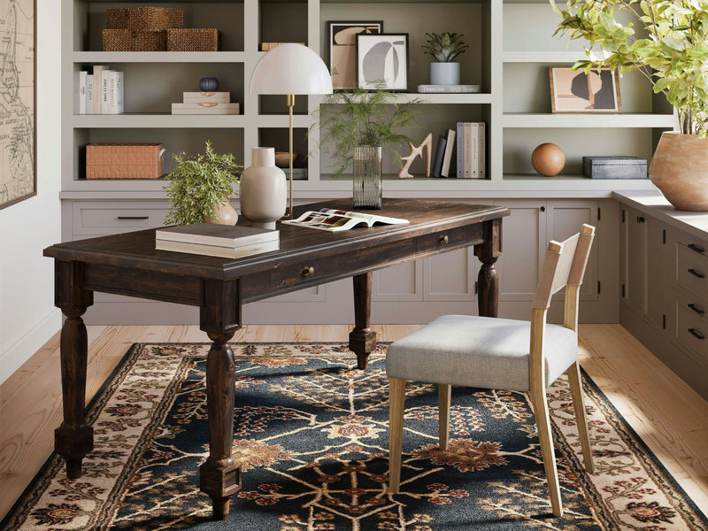 Create a stylish home office with the James+James Rosemary Desk in Tobacco, a cushioned chair, and open shelving in the background. Decorate the desk with books, a plant, and a lamp, while placing various decorative items on the shelves. Add a patterned rug beneath the desk and chair to complete the look.