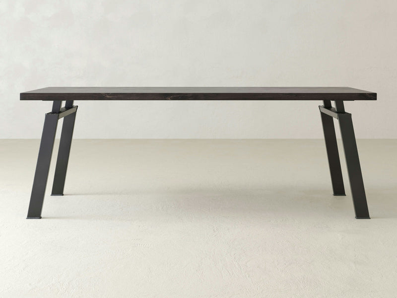 The Fulton Desk - Charred Ember by James+James is a minimalist piece featuring a rectangular wooden top and black metal legs. The sturdy legs are angled outward, lending an industrial aesthetic. Positioned against a plain, light-colored background, the desk's modern design stands out in its simplicity.