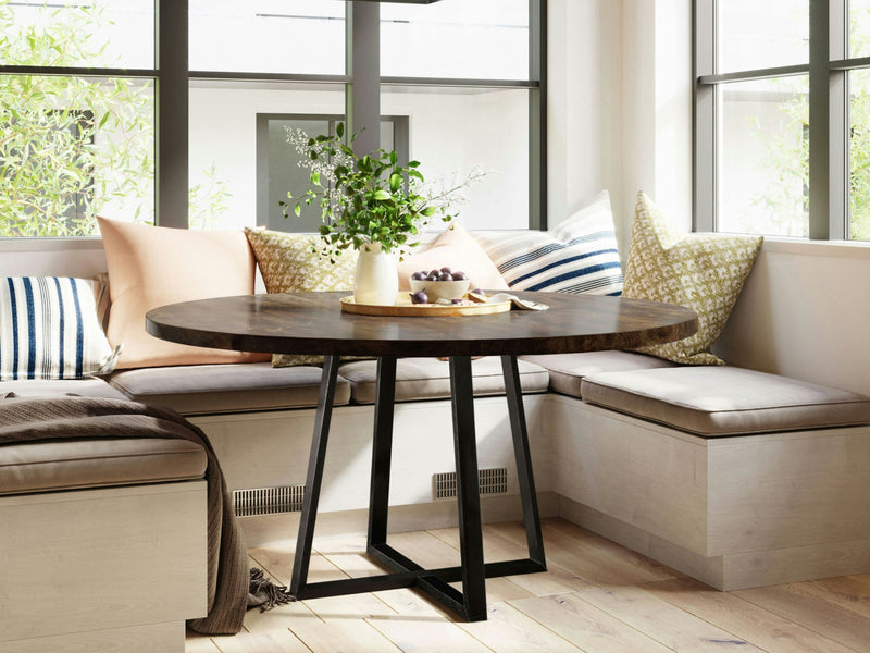 A cozy breakfast nook featuring the Watson Round Dining Table in Tobacco by James+James, with sleek black metal legs and built-in seating adorned with various patterned pillows. A vase filled with green foliage and a fruit bowl rest on the table, while large windows in the background allow natural light to flood the space.