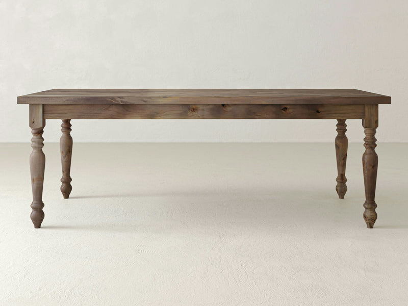 The French Country Dining Table - Barn Wood from James+James features a rectangular top and four intricately carved legs. The wood boasts a natural, rustic finish, and the table is set against a plain, light-colored background.