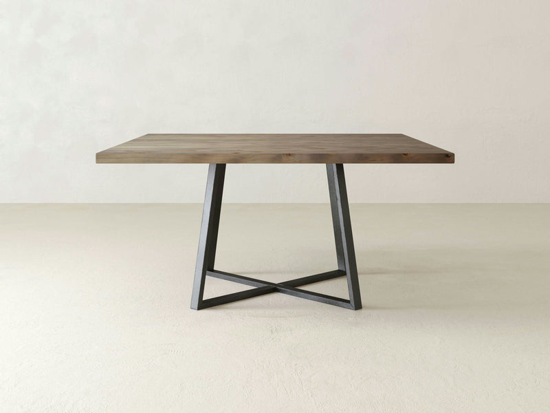 The Watson Square Dining Table by James+James features a minimalist design with a barn wood surface supported by a sleek, geometric metal base. It stands elegantly in a simple, empty room with a light-colored floor and an off-white wall.