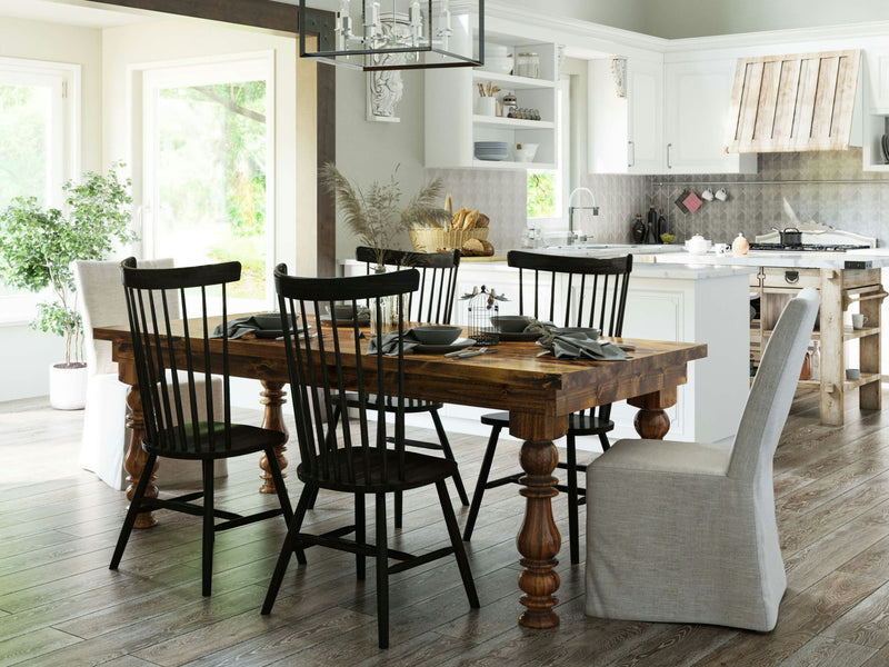 A well-lit kitchen and dining area featuring the Annli Dining Table - Tuscany by James+James, surrounded by a mix of black spindle-back chairs and upholstered chairs. The space includes light-colored cabinets, modern appliances, greenery in a pot, and decorative items on shelves and counters.