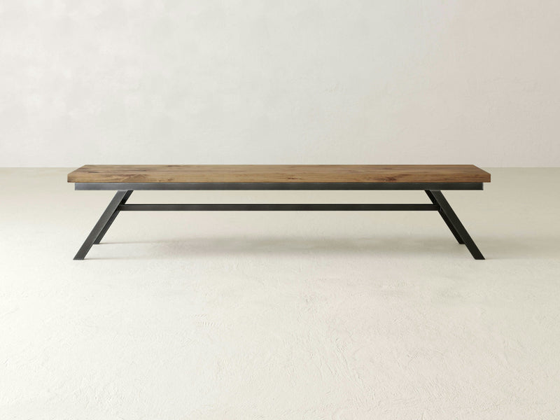 The Fulton Bench - Harvest Wheat by James+James is set against a plain, off-white background. This minimalist wooden bench features a rustic, natural finish and has a single plank seat supported by black, angular metallic legs. The design is simple and modern, emphasizing clean lines and functionality.