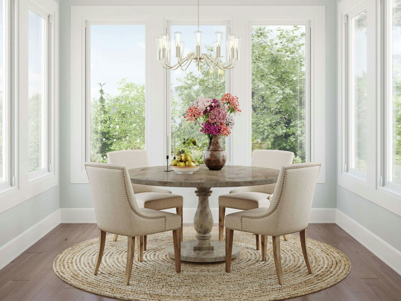 A cozy dining area features the Vivien Round Dining Table – Barn Wood by James+James, adorned with a vase of colorful flowers in the center, surrounded by four beige upholstered chairs. The space is well-lit by large windows and a chandelier, and a woven circular rug lies beneath the table.