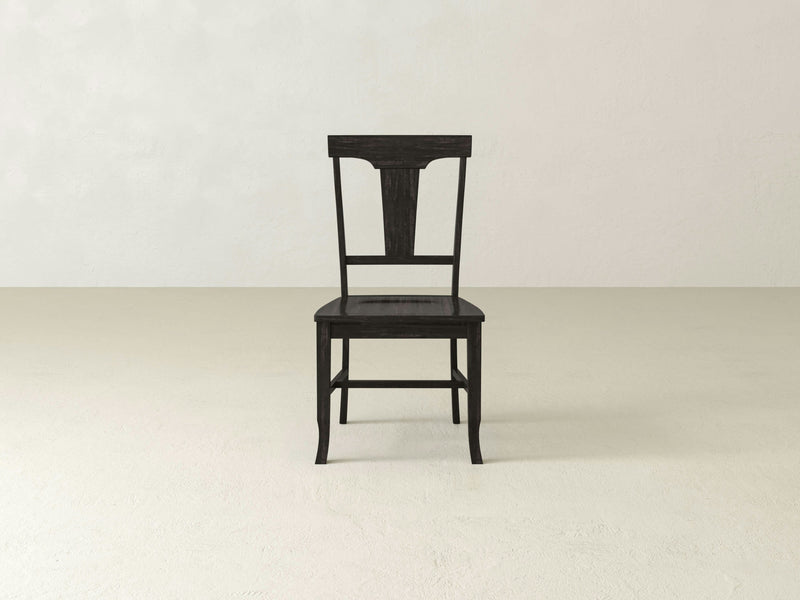 The Grace Dining Chair - Charred Ember by James+James is viewed at an angle, revealing its back and one side. This wooden chair boasts a simple yet elegant design with a rectangular backrest that features a carved top and a flat seat. The legs are slightly curved, enhancing the chair's refined appearance.