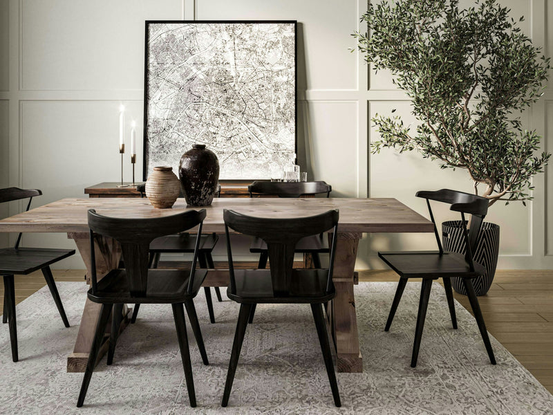 A modern dining room features a Trestle Dining Table in barn wood by James+James, surrounded by six black chairs. On the table are two ceramic vases and a lit candle. A large framed abstract artwork rests on the table against a paneled wall, while an olive tree in a black pot stands beside the table.