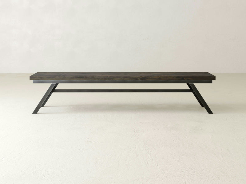 The Fulton Bench - Deep Grey by James+James features a simple design with a flat rectangular top and angled legs, set against a plain off-white background. The bench boasts a minimalist aesthetic characterized by clean lines and a dark finish.