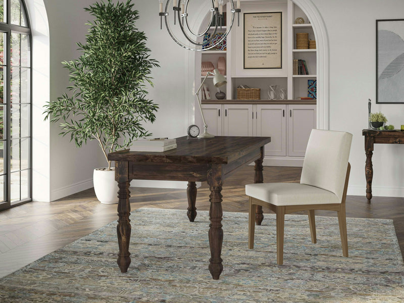 A minimalist home office features the Turned Leg Desk - Tobacco by James+James and a beige upholstered chair. The room has a large green potted plant, a chandelier, built-in white bookshelves with decor, and a light-colored rug on a polished wooden floor. Archways and windows allow natural light in.