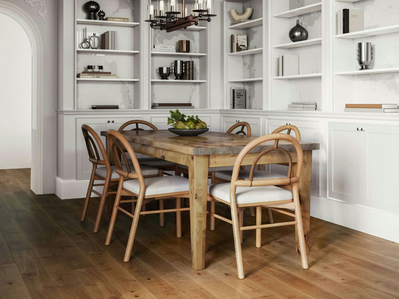 A cozy dining room featuring the Ava Parsons Dining Table in Harvest Wheat by James+James and six chairs with curved wooden backs and cushioned seats. The room showcases white built-in bookshelves adorned with books, decorations, and potted plants, all set against a floor made of wooden planks.