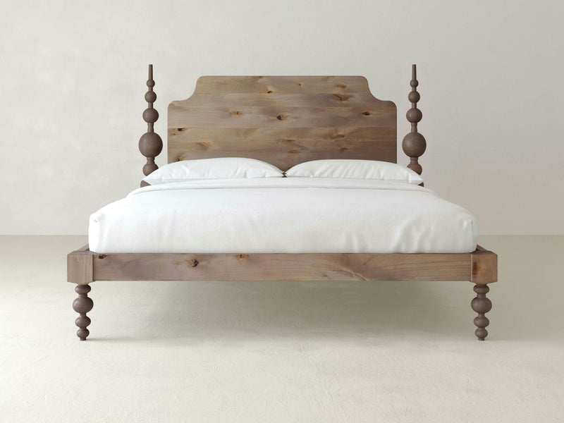 The Opal Bed - Barn Wood by James+James features intricately carved posts supporting a neatly made bed with white bedding and pillows. The headboard showcases a smooth, rustic design. This elegant bed is placed in a minimalist room with white walls and a light-colored floor.