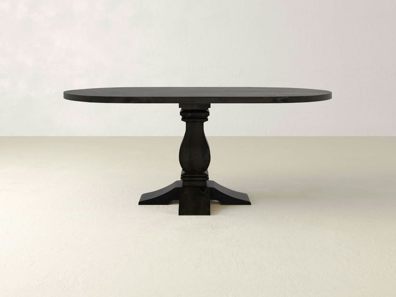 A dark wooden oval dining table, known as the Heirloom Oval Dining Table - Charred Ember by James+James, stands against a plain, light-colored background. The table features a smooth, polished surface and a single ornate pedestal base that gives it a sturdy appearance.