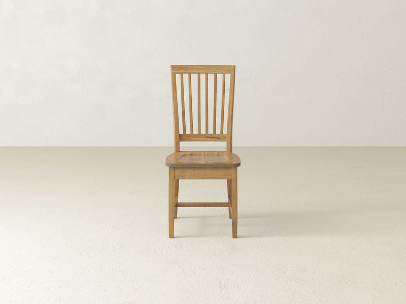 The Jane Dining Chair - Harvest Wheat by James+James is a wooden chair with a natural finish. It features a high, slatted backrest and a solid seat. The chair has four slightly curved legs connected by support bars near the bottom, offering simple and classic design perfect for dining or casual seating.