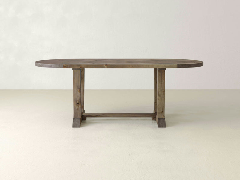 A Thaden Oval Dining Table - Barn Wood by James+James, featuring an oval-shaped top and two sturdy legs connected by a horizontal support bar, graces an empty room with light-colored walls and floors. The table boasts a rustic, natural finish.