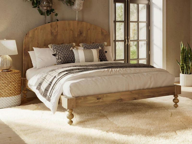 A cozy bedroom features the Eden Bed in Harvest Wheat by James+James, adorned with white and patterned pillows and a beige blanket. The room includes a soft, light-colored rug, a wicker side table with a lamp, large windows letting in abundant natural light, and a green potted plant in the corner.