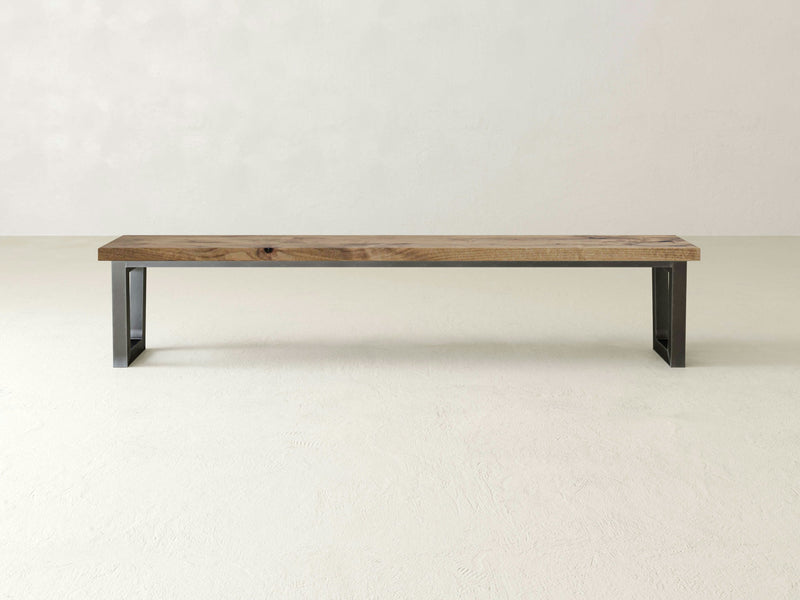 The Trapezoid Bench by James+James, featuring a solid hardwood top supported by two trapezoidal industrial steel bases, is placed against a plain, light-colored wall on a matching floor, highlighting its simple yet elegant design.