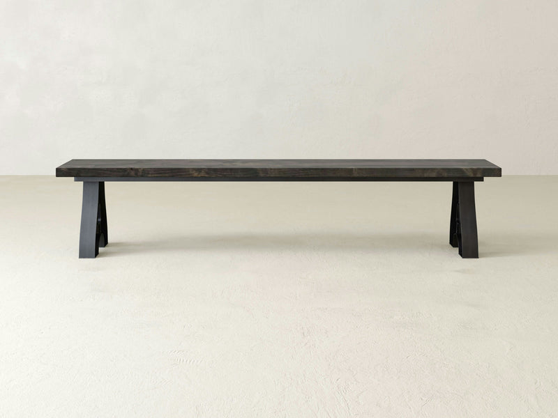 The Wishbone Bench - Deep Grey by James+James stands against a plain, light-colored background. This minimalist wooden bench features a simple, rectangular seat with straight, angular legs, creating a modern aesthetic.