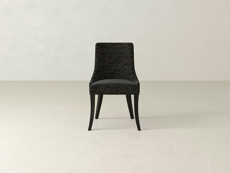 A single James+James Willow Dining Chair, upholstered in dark grey with tapered black legs, sits on a plain light-colored floor against a light grey wall. The chair, featuring a slightly curved backrest and plush padded seat, adds timeless elegance to the minimalist setting.