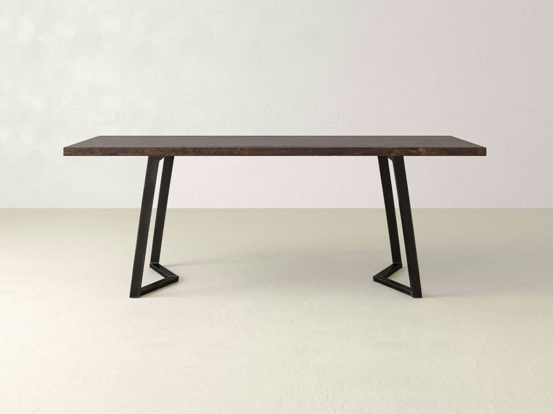 The Arkwright Dining Table - Tobacco from James+James is a minimalist piece featuring a dark brown rectangular wooden top and black metal legs arranged in a geometric design. The table is showcased in an empty room with light grey walls and an uncluttered floor.