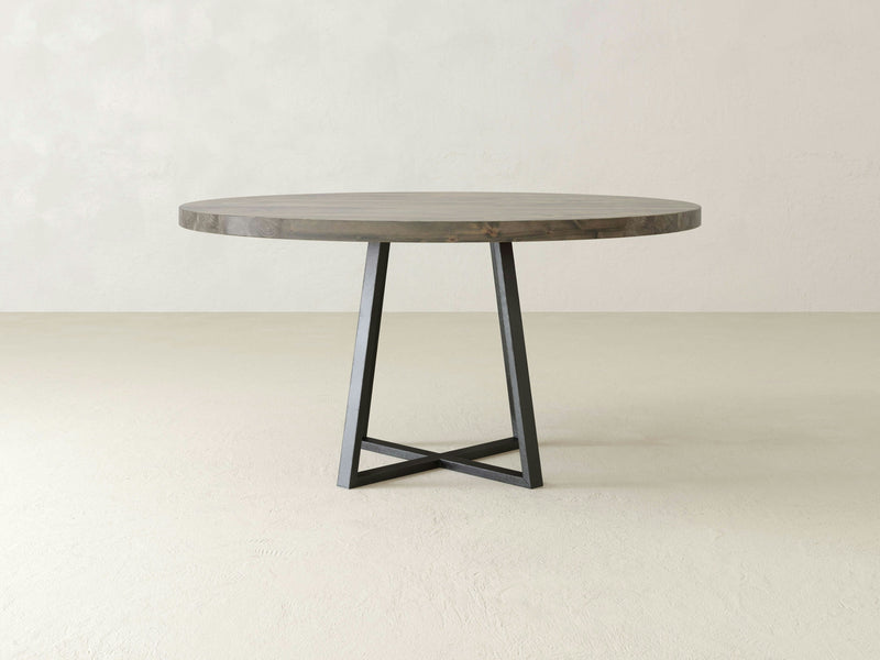 The Watson Round Dining Table - Deep Grey by James+James features a smooth, light-colored top paired with dark, angled metal legs, set against a plain off-white background. Its minimalist and modern design showcases clean lines and a simple, functional aesthetic.