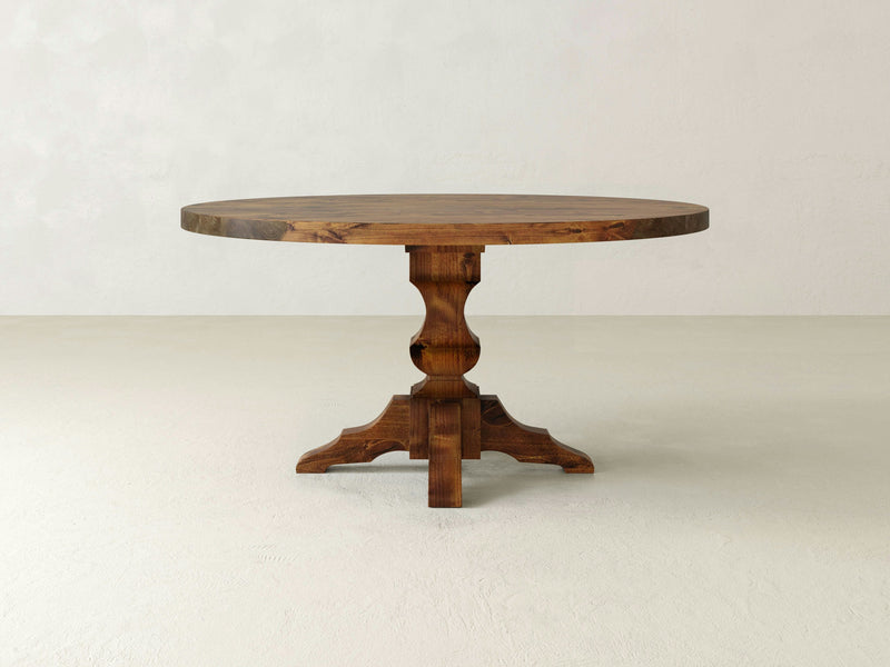 The James+James Sybil Round Dining Table features a thick, flat top and a single, sturdy pedestal base with four curved legs. Handcrafted from hardwood, this table boasts a rustic, natural wood finish and serves as the perfect centerpiece dining table against a plain, light-colored background.