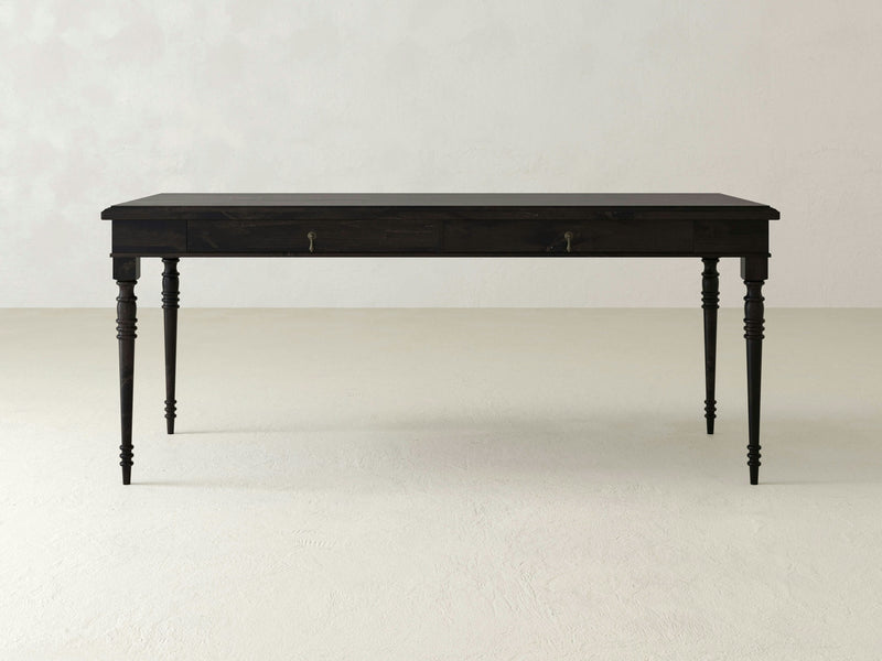 A striking Audrey Desk - Charred Ember from James+James, showcasing a dark wooden finish and turned legs, with two small drawers on either side. Set against a plain light-colored background, this desk embodies a simplistic and classic design while standing confidently on a flat surface.