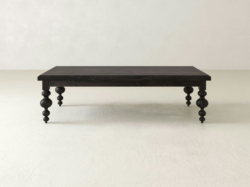 The Olivia Coffee Table - Charred Ember by James+James boasts a rectangular top made of dark wood, supported by ornately turned legs. Set against a plain, light-colored backdrop, this minimalist table strikes a perfect balance between simplicity and elegance with its intricate leg details.