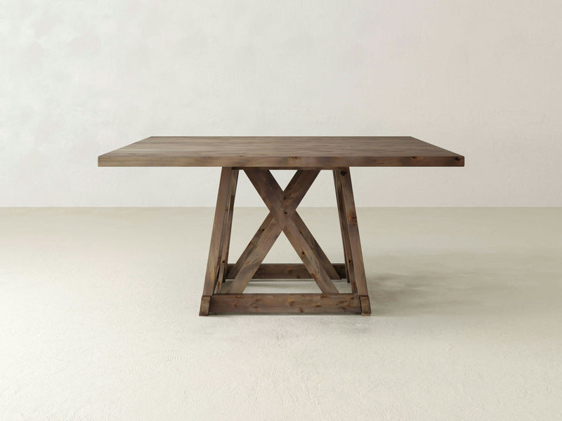 The Julia Square Dining Table by James+James is crafted from barn wood, featuring a square top and a base with a cross-braced design. Positioned against a pale, neutral-colored background, the table showcases its natural finish, emphasizing the grain and texture of the wood.