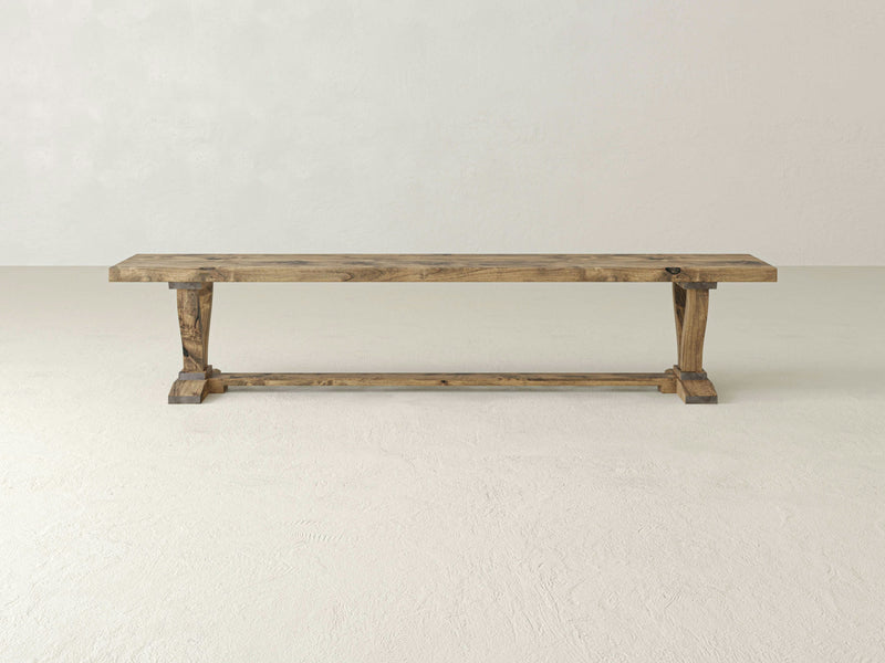 The Vera Bench - Harvest Wheat by James+James is a long, rustic wooden bench featuring a natural finish and X-shaped supports at both ends, connected by a horizontal wooden stretcher. It appears sturdy and is suitable for both indoor and outdoor use, showcasing a simple yet elegant design.