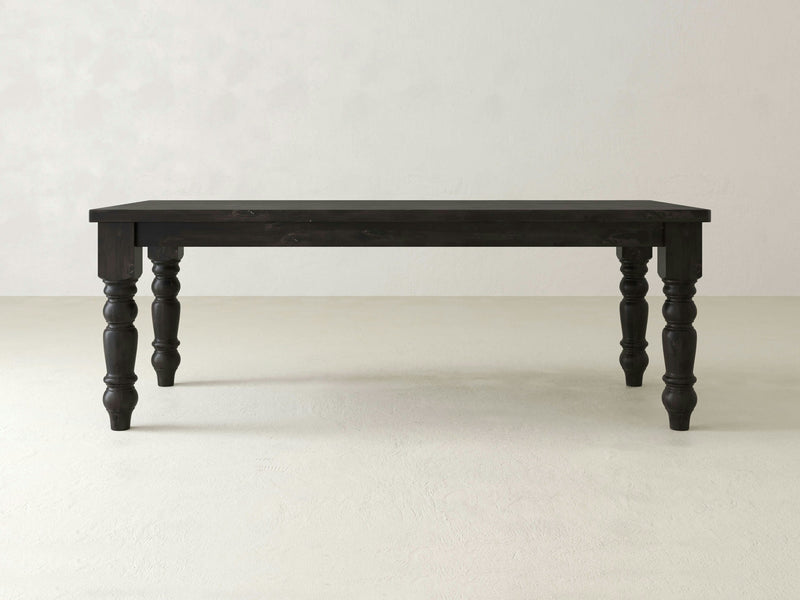 A James+James Baluster Dining Table in the Charred Ember finish, featuring four ornately carved legs, stands on a light-colored floor against a plain, light-colored wall. The table has a rectangular shape and a smooth surface. The overall design is classic and elegant.