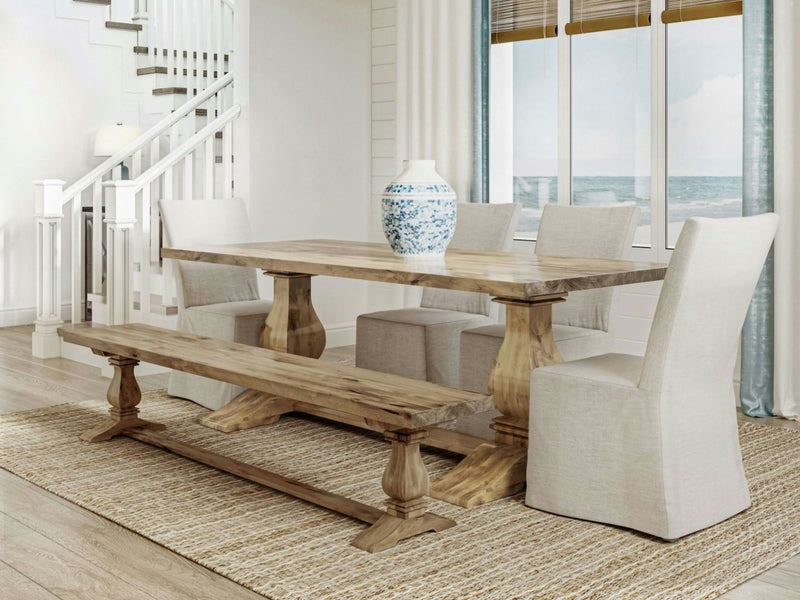 A dining room with a rustic wooden table and a matching Heirloom Bench in Harvest Wheat from James+James. The table is accompanied by four beige upholstered chairs. A large white and blue ceramic vase is centered on the table. The room features a woven rug and has a view of the ocean through large windows.