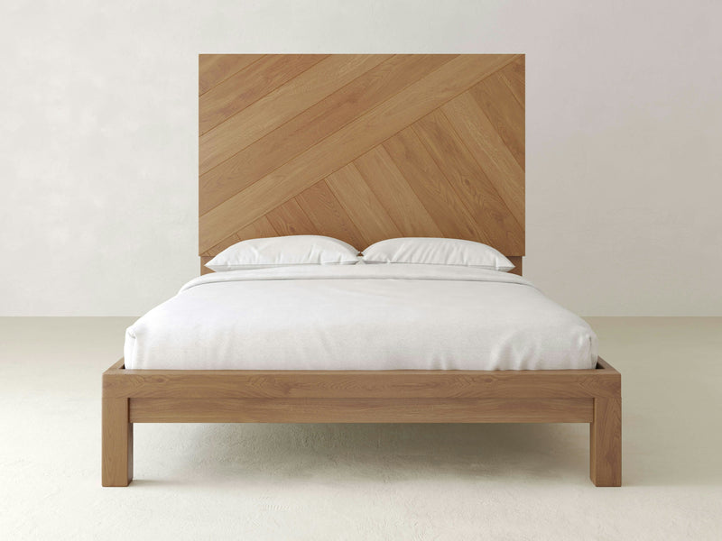 Introducing the Luna Bed by James+James: a minimalist masterpiece made from natural white oak, showcasing a tall headboard adorned with an elegant chevron pattern. The bed is perfectly complemented by crisp white bedding and pillows, set against light-colored walls and flooring that beautifully accentuate the natural wood finish of this refined piece.