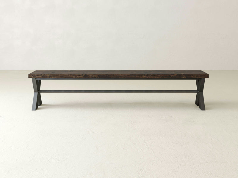 The X-Base Bench - Tobacco by James+James, a sleek black wooden bench with a minimalist design, is positioned against a plain, light-colored background. The bench features a long seat atop two angular legs connected by a horizontal support bar.