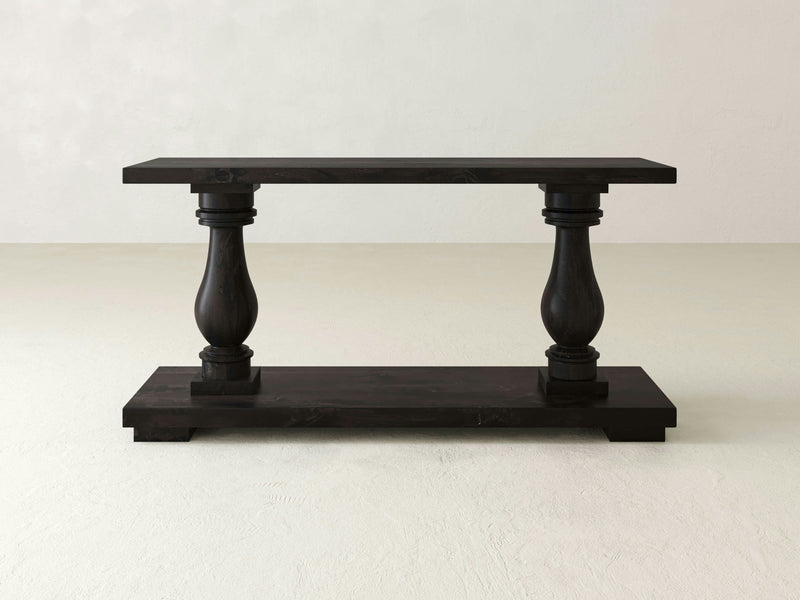 The Vivien Sofa Table - Charred Ember by James+James features a dark wood finish with two ornate, turned legs. The table has a flat rectangular top and a wide base, all set against a plain, light-colored background.