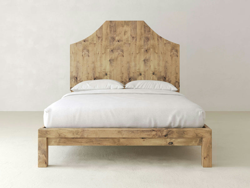 The Megan Bed - Harvest Wheat by James+James, featuring a minimalist wooden design and a tall, natural wood headboard, sits against a plain white wall. The bed is made up with white bedding and two pillows, creating a simple and clean aesthetic. The wood grain of the headboard and frame is clearly visible.