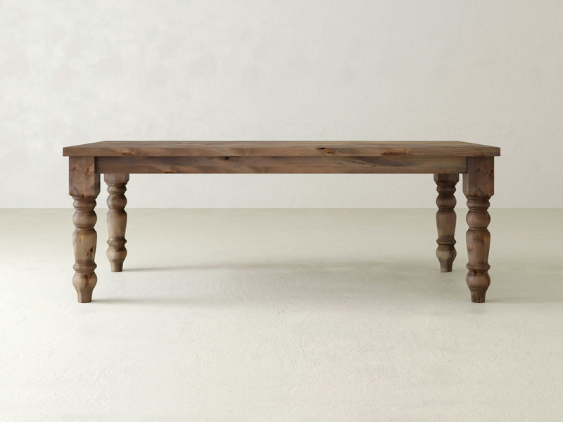 A Baluster Dining Table - Barn Wood from James+James, with a rectangular top and four intricately carved spindle legs, placed in an empty, light-colored room. The table features a natural, rustic finish that highlights the beautiful wood grain and knots.