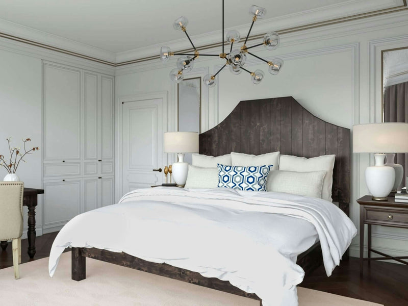 A modern bedroom featuring the James+James Megan Bed in Charred Ember, adorned with white bedding and blue and white accent pillows. The room has light-colored walls with decorative molding, a contemporary chandelier, bedside tables with lamps, and a small desk with a chair in the corner.