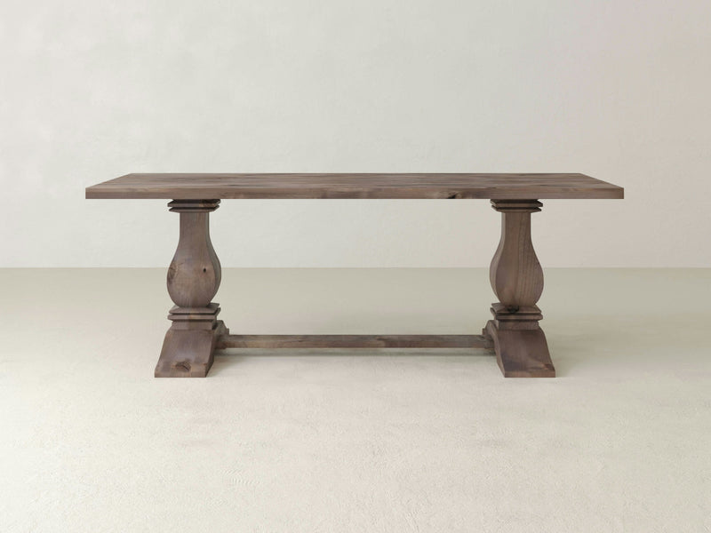 The Heirloom Dining Table - Barn Wood by James+James showcases a rectangular wooden top, supported by two ornate pedestal legs connected with a horizontal support beam. It features a rustic finish and is displayed on a light-colored floor against a plain, light-colored wall.