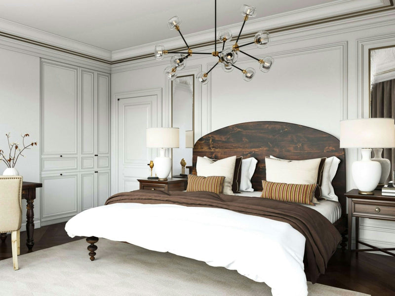 Elegant bedroom featuring the Eden Bed in Tobacco by James+James with a dark wooden headboard and white bedding with brown accents. The room has two matching bedside tables with lamps, a modern chandelier, white paneled walls, and a desk with a chair and decorative vase in the corner.